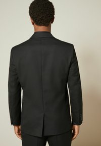 Veste de costume noire avec un design structuré, présentant une fente arrière, des revers crantés et un tissu texturé. Manches avec accents de boutons.