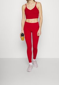 Červený sportovní crop top a odpovídající legíny s vysokým pasem, hladký materiál. Drží žlutou láhev na nápoje, má na sobě světle růžové sportovní boty.