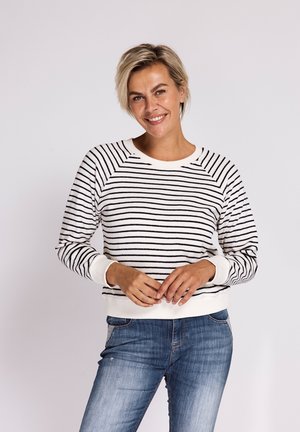 Lächelnde Frau mit kurzen blonden Haaren, die einen weißen Pullover mit schwarzen horizontalen Streifen und blaue Jeans trägt, posiert mit verschränkten Händen.
