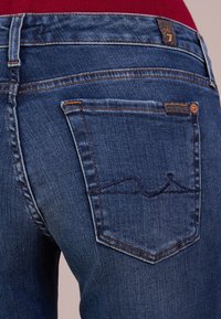 Denimjeans i medelblått med en slät yta. Har två bakfickor, varav en har ett sömnt mönster i v ågor och orangea stickningar.