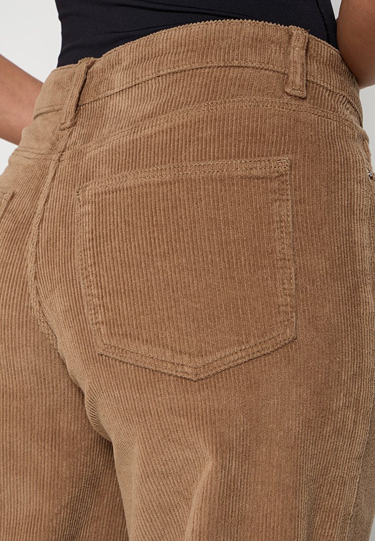 Pantaloni di velluto a coste marroni con una superficie testurizzata e rigature visibili, caratterizzati da una tasca a toppa sul retro e un design a vita alta.