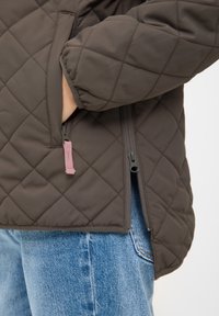 Hand in de zak van een gequilt bruin jack met een roze ritssluiting, gecombineerd met blauwe denim jeans.