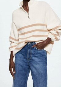 Pull crème à col, avec des rayures beige horizontales et une texture côtelée. Associé à un jean bleu taille haute et coupe droite.