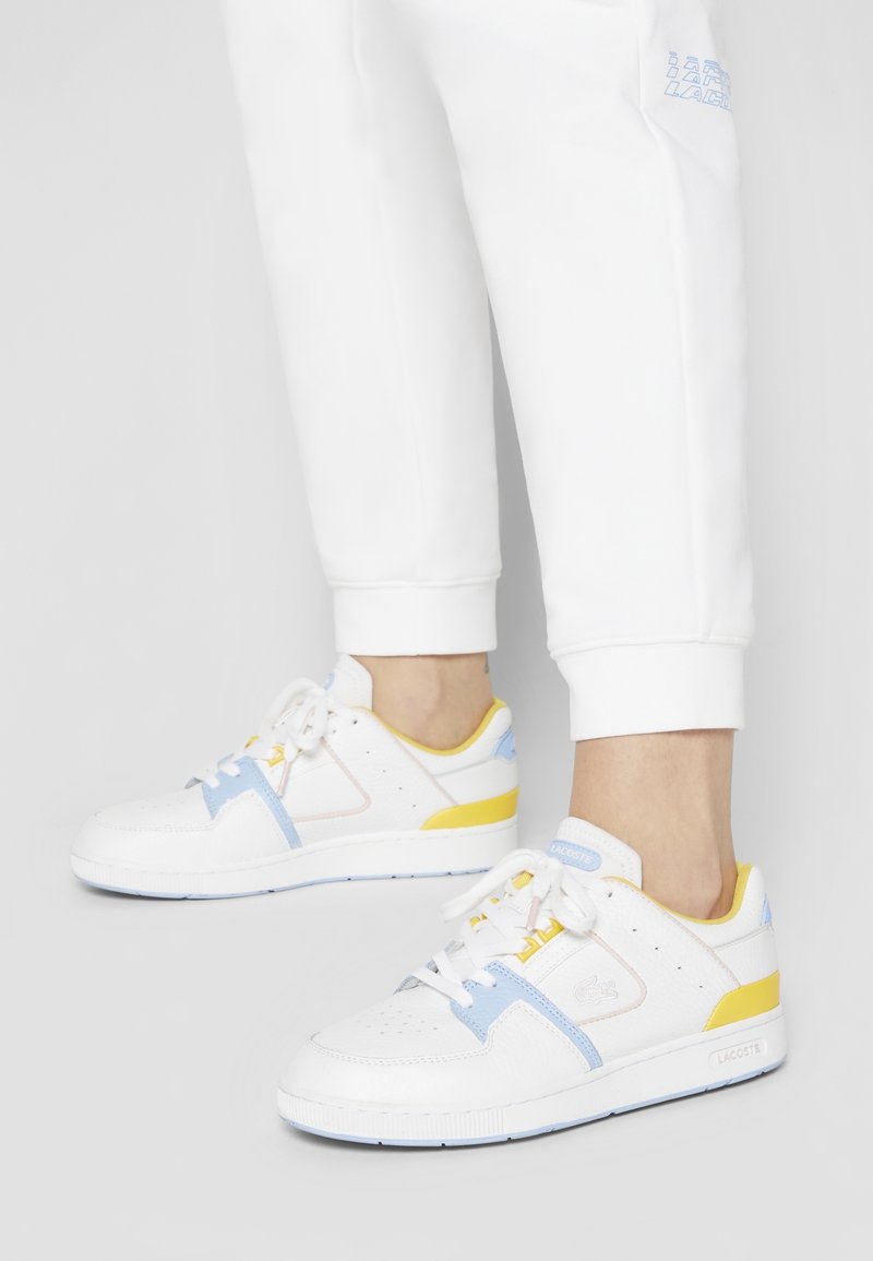 Baskets blanches avec des accents pastel bleu, jaune et rose. Tige en cuir texturé, bout perforé et semelle en caoutchouc. Marque LACOSTE visible.