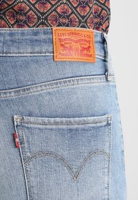 Jeans de denim de color azul claro con una etiqueta de marca de cuero con detalles en rojo y dorado, y costura clásica en los bolsillos traseros con un diseño curvado.