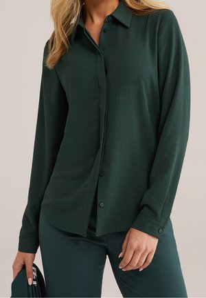 Kvinde iført mørkegrøn knap-op skjorte og matchende bukser, der holder en mørkegrøn genstand i sin venstre hånd.