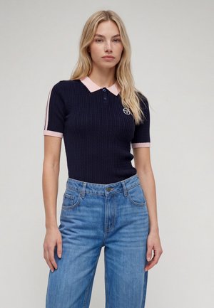 Femme aux longs cheveux blonds portant un polo tricoté bleu marine à manches courtes avec col et poignets roses, et un jean bleu sur fond uni.