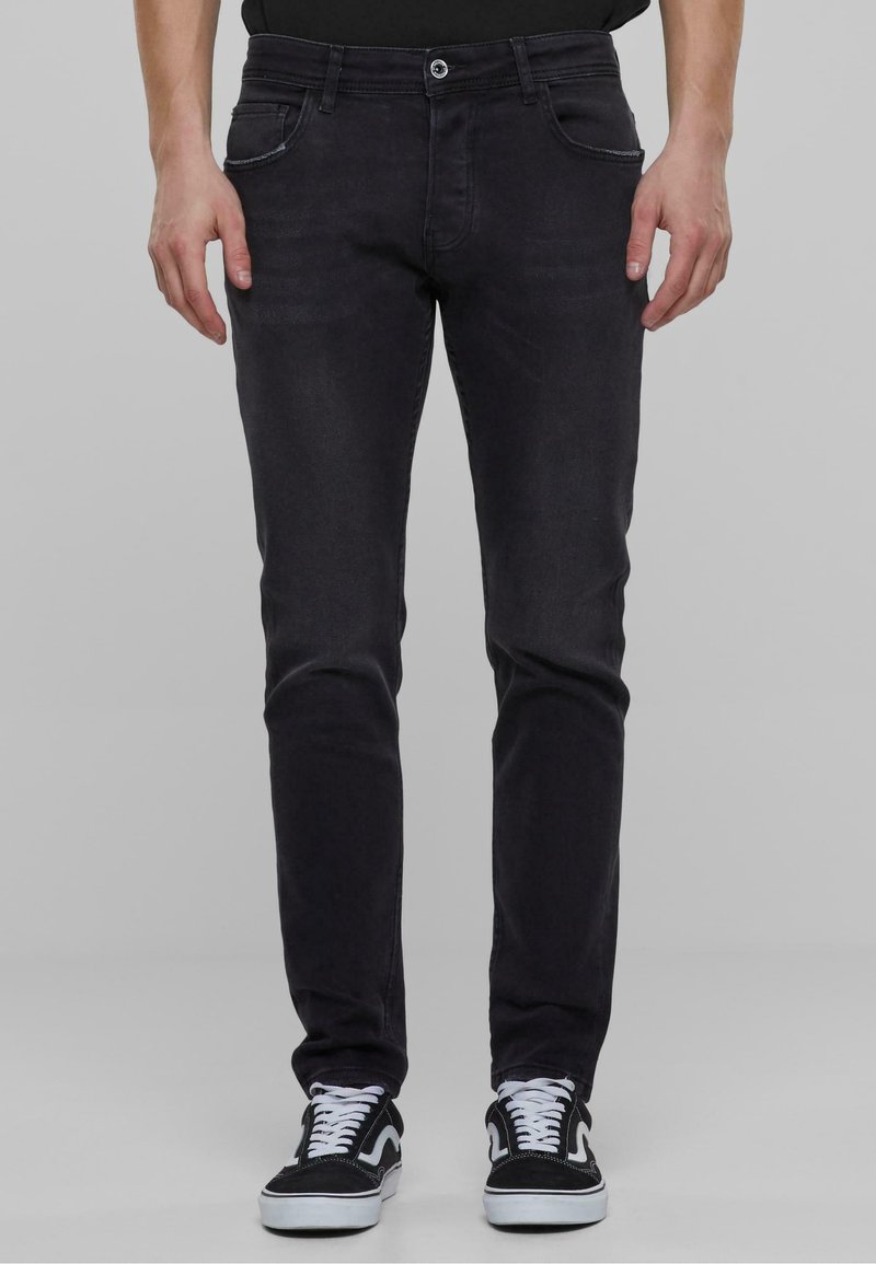 Crne traperice slim-fit kroja s glatkom teksturom, pet džepova i standardnim zatvaranjem na dugme, uparenih s crno-bijelim tenisicama.