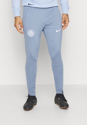 Mann trägt hellblaue Nike-Trainingshose mit Inter-Mailand-Logo und schwarze Nike-Fußballschuhe, steht mit entspannten Händen an den Seiten.