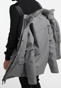 Giacca esterna grigia con una finitura testurizzata, dotata di una doppia cerniera frontale e tasche a patta, tenuta aperta su uno sfondo di maglione nero.