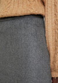 Lähedalt vaade isikule, kes kannab pruuni kudumist kampsunit ja kõrge vöökohtadega halli villast seelikut, tuues esile tekstuurid ja kangaste detailid.