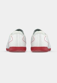 Zapatillas deportivas blancas con un upper texturizado, acentos rojos en la suela y en el lateral, y un acabado liso. La vista trasera muestra costuras distintas y detalles de diseño.
