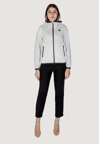 Giacca a vento bianca con cappuccio, dettagli neri e tasche con zip, abbinata a pantaloni neri e tacchi beige.