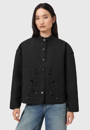 Giacca nera trapuntata con colletto alla mandarino, caratterizzata da intagli floreali e bottoni a scatto, abbinata a jeans in denim blu chiaro.
