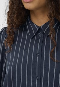 Nāvpulēta zila pogājamā kreklā ar vertikālām baltām svītrām, teksturētu audumu, spetsētu apkakli un divām redzamām pogām pie apkakles.