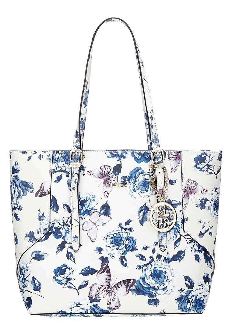 Valkoinen kassi, jossa on sininen kukkakuosi ja violetteja perhosia, kaksinkertaiset kahvat, kultasävyinen logokoru ja Guess-brändäys etupuolella.