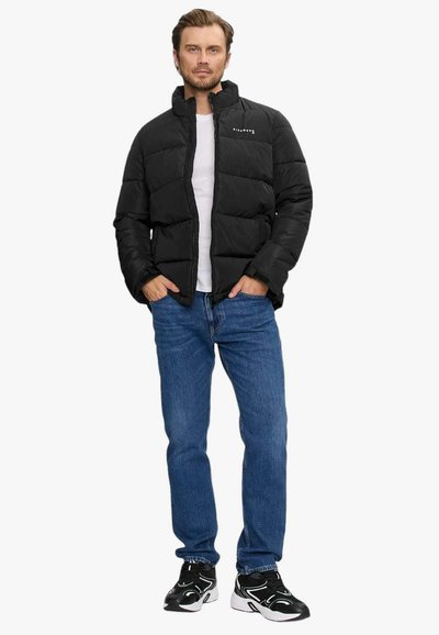 Giubbotto puffer nero con colletto alto, chiusura frontale con zip e dettagli del logo, abbinato a jeans blu e sneakers nere. Tessuto dal tocco liscio.
