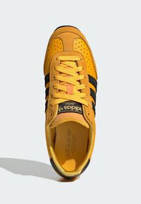 Gelbe Wildleder-Sneaker mit schwarzen Lederstreifen, perforierter Zehenpartie, flachen Schnürsenkeln und gepolstertem Schaft. Branding auf der Zunge und der Innensohle.