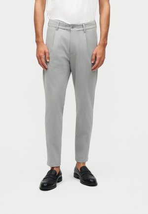 Man draagt lichtgrijze, op maat gemaakte broek en zwarte leren loafers, staand tegen een witte achtergrond.