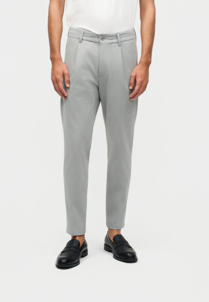 Mann iført lysegrå skreddersydde bukser og svarte skinnloafers, stående mot en hvit bakgrunn.