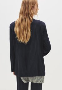 Tengerészkék csíkos blazer, testre szabott fazonnal, sima textúrával és látható varrásokkal, szürke, bolyhos alsó résszel párosítva.