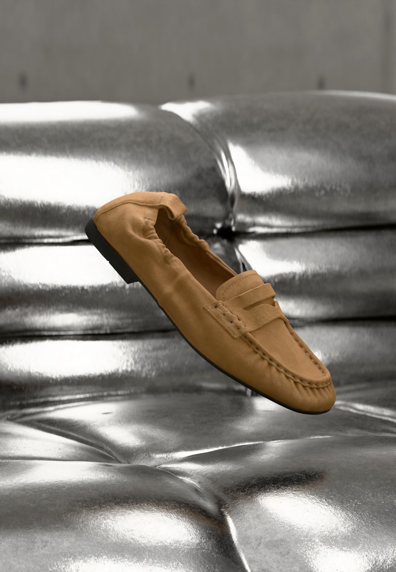 Chaussure mocassin en daim beige flottant au-dessus d'un canapé en cuir argenté brillant et capitonné.