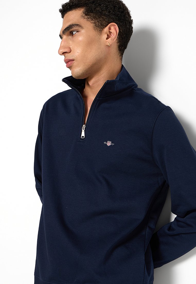 Navyblauer Reißverschluss-Pullover aus weichem Stoff, mit hohem Kragen und kleinem Logo auf der Brust. Glatte Textur mit lässiger Passform.