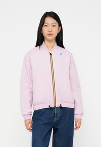 Jeune femme portant une veste zippée rose clair avec un détail de zip rayé et un jean bleu, se tenant contre un fond blanc.