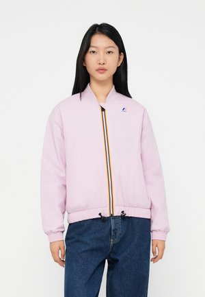 Jeune femme portant une veste zippée rose clair avec un détail de zip rayé et un jean bleu, se tenant contre un fond blanc.