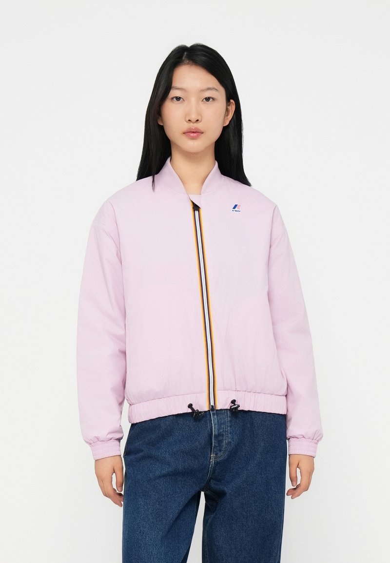 Jeune femme portant une veste zippée rose clair avec un détail de zip rayé et un jean bleu, se tenant contre un fond blanc.