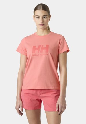 Mujer con camiseta Helly Hansen color coral y pantalones cortos a juego, de pie frente a un fondo liso y claro.
