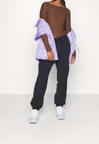 Žena v černých teplácích, bílých teniskách a hnědém topu s dlouhým rukávem, která drží levandulovou péřovku přehozenou přes ruce.