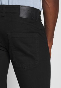 Jeans de mezclilla negra con un parche de cuero que lleva el nombre de la marca "INDICODE". Detalles de bolsillos estructurados con costuras en contraste para un diseño adicional.