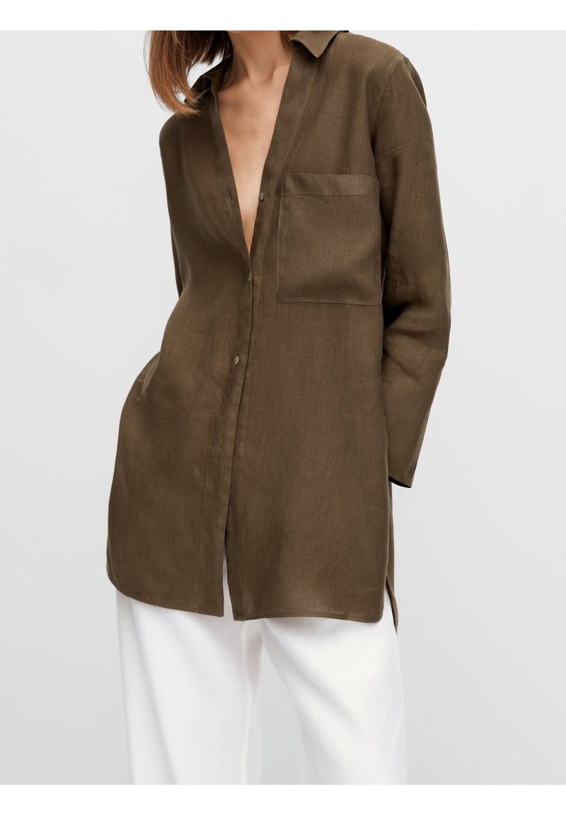 Femme portant une chemise à manches longues vert olive avec une poche avant et un pantalon blanc ample, debout devant un fond clair uni.