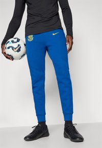Calças de treino azuis da Nike com um logótipo amarelo e o emblema do Barcelona, apresentam um corte ajustado e punhos em côtel, combinadas com sapatilhas de desporto pretas.