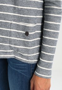 Randig grå och vit långärmad topp med texturerat tyg, sidoficka och metallisk logotypdetalj nära fållen, kombinerad med blå denim.