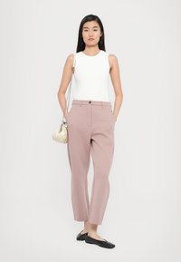 Haut blanc sans manches ajusté, pantalon rose clair élégant avec poches latérales, et chaussures plates noires. Portant un petit sac à main crème.