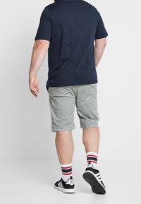Mies, joka wearing tummansinistä t-paitaa, vaaleanharmaita polvipituisia shortseja, valkoisia sukkia, joissa on punaiset ja tummansiniset raidat, sekä musta-valkoisia Adidas-urheilukenkiä, kävelee pois.