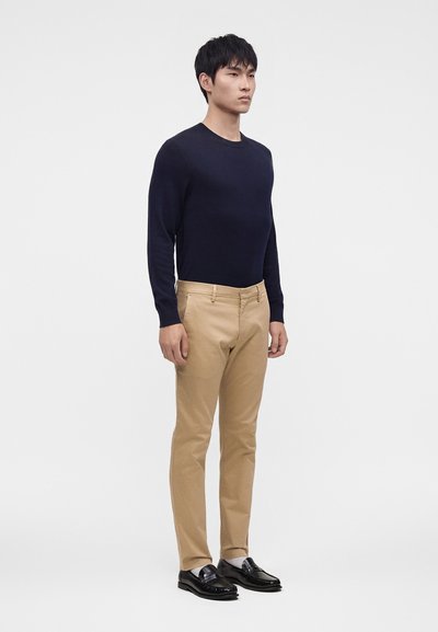 Mand står iført en sort langærmet skjorte, beige bukser, hvide sokker og sorte loafers på en ensfarvet hvid baggrund.