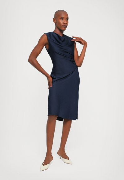 Rochie de satin albastru navy cu decolteu drapat, fără mâneci, și tăietură până la genunchi. Purtați cu mule albe cu toc, evidențiind o textură netedă.