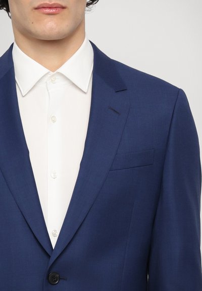 Veste de costume bleu marine avec une coupe ajustée, revers cranté et fermeture à un bouton, associée à une chemise habillée blanche avec un col pointu.