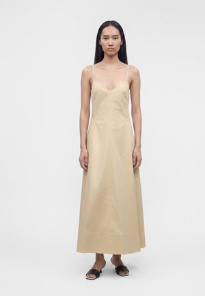 Kvinde med langt, glat hår iført en beige ærmeløs V-hals kjole, der går til anklerne, og sorte flade sandaler med åbne tæer, stående foran en ensfarvet baggrund.