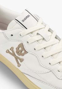 Zapatillas de cuero blancas con un diseño de calavera y tibias en color marrón, suela texturizada amarilla y cordones crema con un logotipo en la lengüeta.