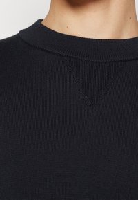 Gros plan sur un pull noir à col rond côtelé avec un détail de couture triangulaire texturée au niveau de l'encolure, porté autour du cou et de la partie supérieure de la poitrine d'une personne.
