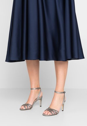 Kvinde iført metallic sølvfarvede strop sandaler med høj hæl og en mørkeblå midi-nederdel mod en ensfarvet baggrund.