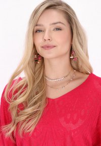 Rosa stickad tröja med ett texturerat mönster. Modellen bär lager på lager av halsband, ett med pärlor, och färgglada hängande örhängen.