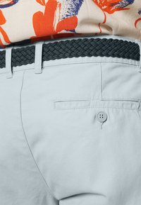 Un pantalon en coton bleu clair présente une ceinture tissée gris foncé et une poche arrière avec une fermeture à bouton. Le design est ajusté et fonctionnel.
