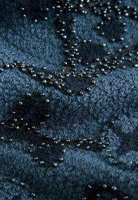 Stickad blåtextil med ett mönster av mörka fläckar och intrikata pärlbroderier, som visar upp varierande texturer och en mjuk yta.