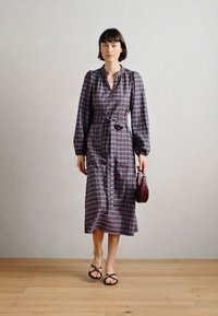 Langs plaidkjole i mørke toner, med V-utringning, lange ermer, innsnevret liv og splitt i siden. Holder en liten burgunder håndveske.