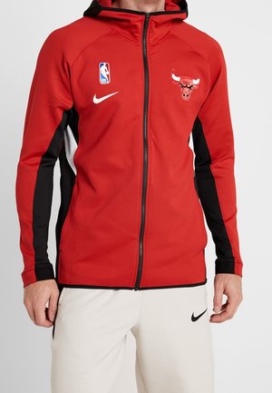 Mężczyzna w czerwonej bluzie z kapturem zapinanej na zamek Chicago Bulls z czarno-białymi akcentami oraz białych spodniach Nike z czarnym logo Swoosh.